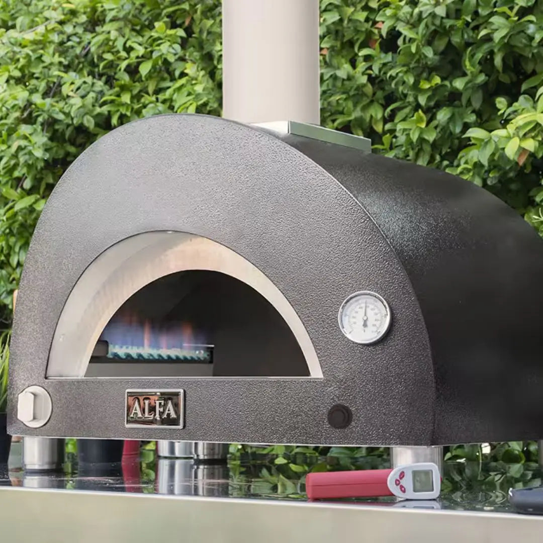 Alfa Classico 2 Pizze Hybrid Gas pizza oven with flames inside, brand 'Alfa' visible, against a green hedge background.