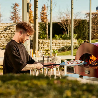 Man preparing pizza in an outdoor setting with the Alfa Moderno 3 Pizze Wood Fired Pizza Oven in Antique Red