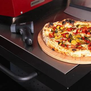 Pizza on a wooden board next to a Alfa pizza oven and a pizza stone.