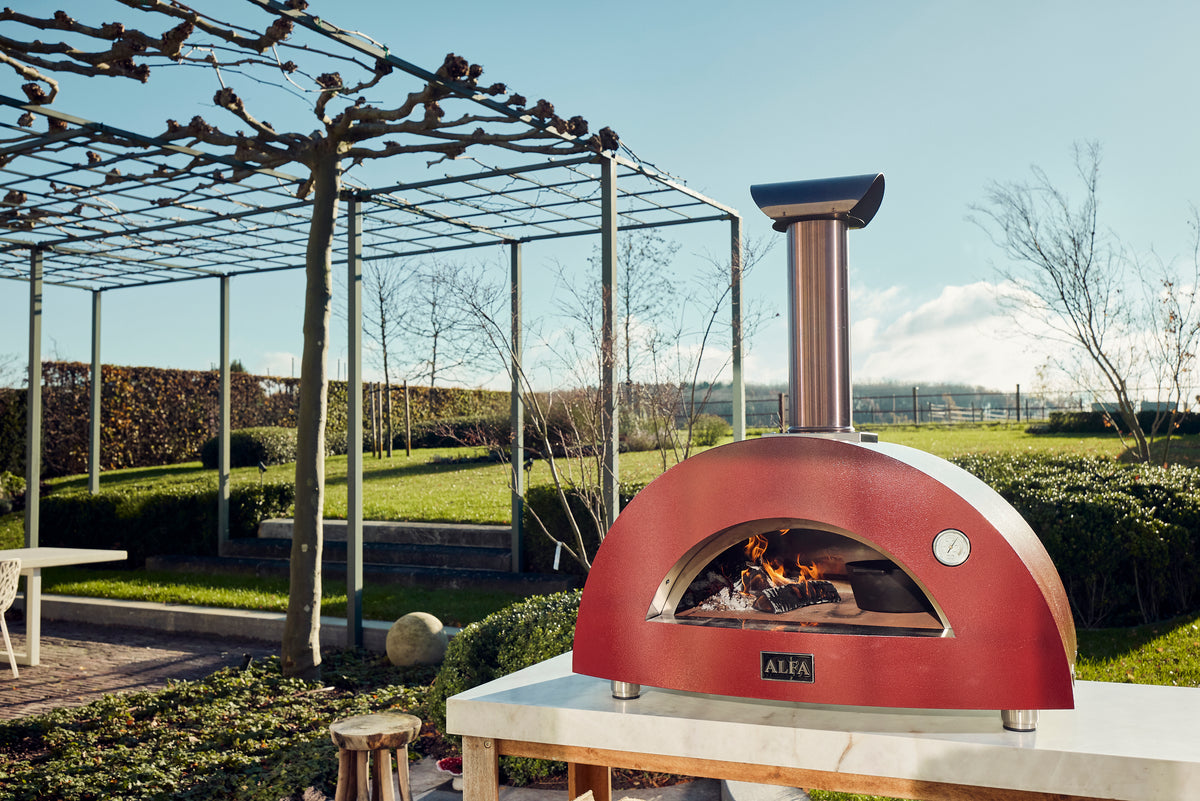 A red Alfa pizza oven, lit, on an outdoor worktop beside a pergola