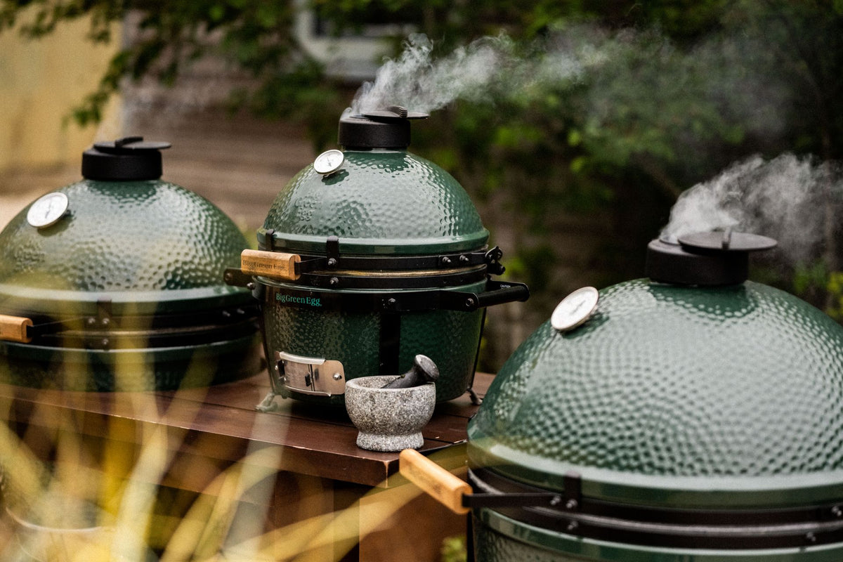 Three Big Green Egg kamado grills with smoke rising from their lids, on a wooden outdoor worktop in a garden
