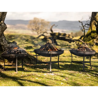 Three Dean Forge Moorland Firebowl fire pits with burning wood in a grassy outdoor setting with trees and mountains in the background.