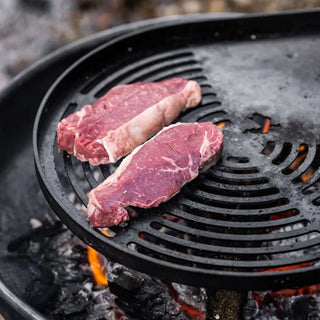 Two raw steaks on Dean Forge Moorland Firebowl Swing Arm grill pan over an open fire.