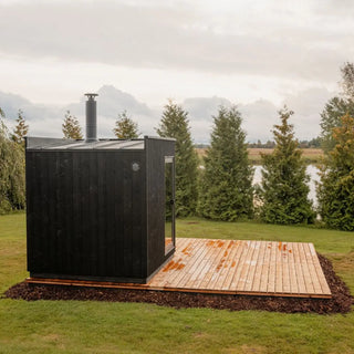 Black outdoor sauna with wooden platform in a scenic landscape with trees and water.