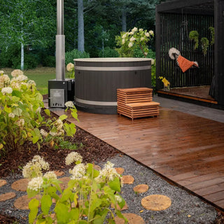 Outdoor hot tub setup with wooden deck, plants, and a small building in the background.