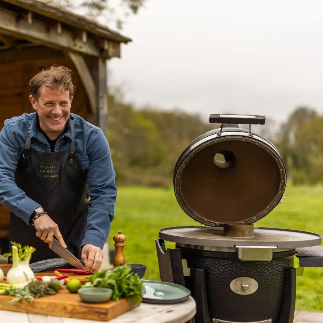Matt Tebbutt preparing food outdoors next to Monolith Avantgarde kamado grill in a garden setting.