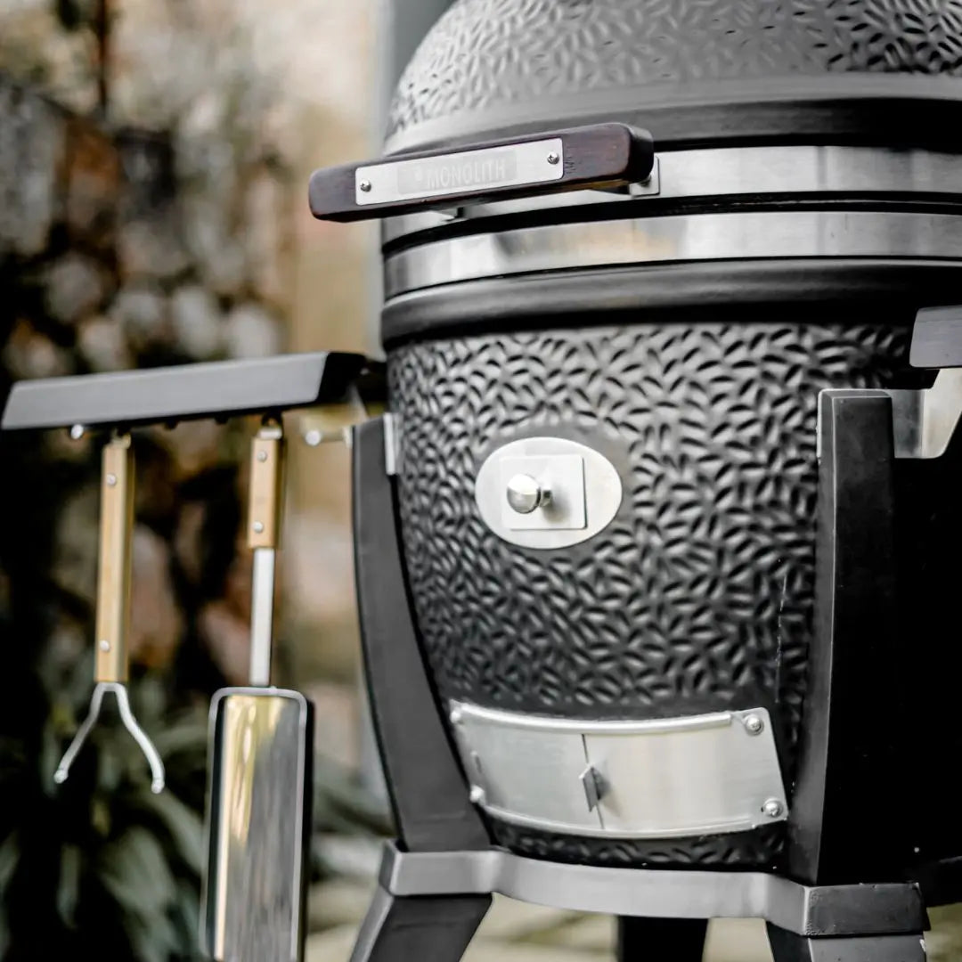 Close-up of a large Monolith Avantgarde kamado grill bbq with tools attached, set against a blurred natural background.