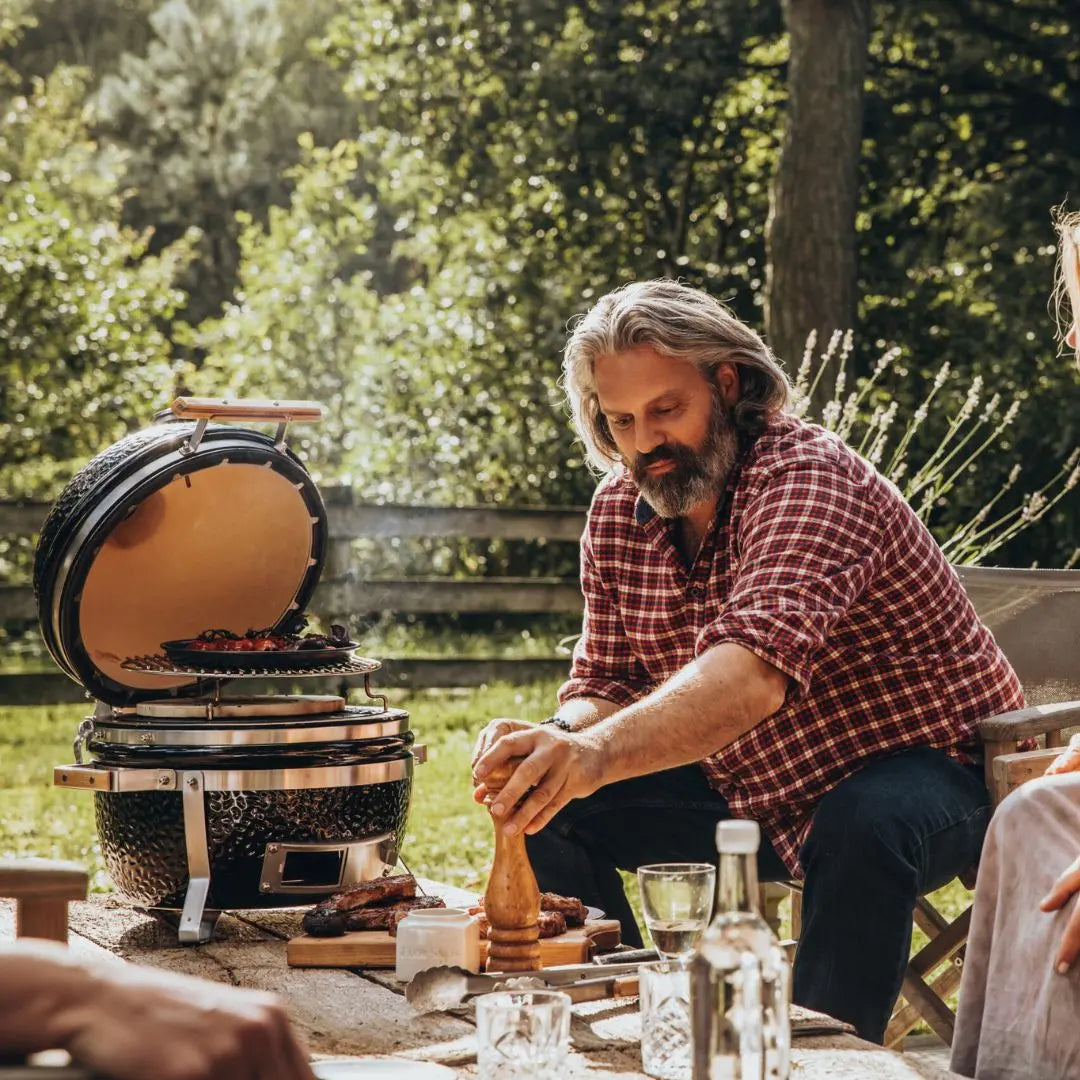 Man seasoning food on a Monolith ICON 53cm black kamado grill outdoors with trees in the background