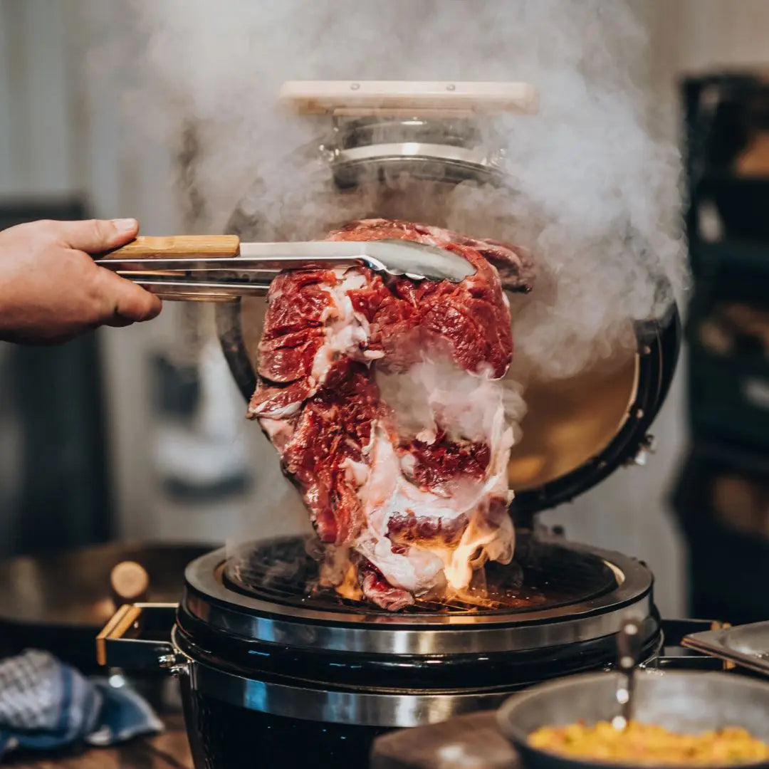 Person grilling a large piece of meat over a flame with smoke rising on Monolith ICON 53cm kamado grill bbq