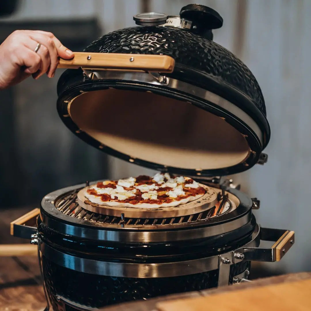 Person opening black Monolith ICON 53cm kamado grill to reveal a pizza inside.