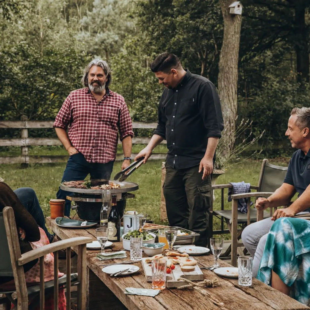 Three men around a wooden table outdoors, one grilling food on Monolith ICON 53cm kamado grill