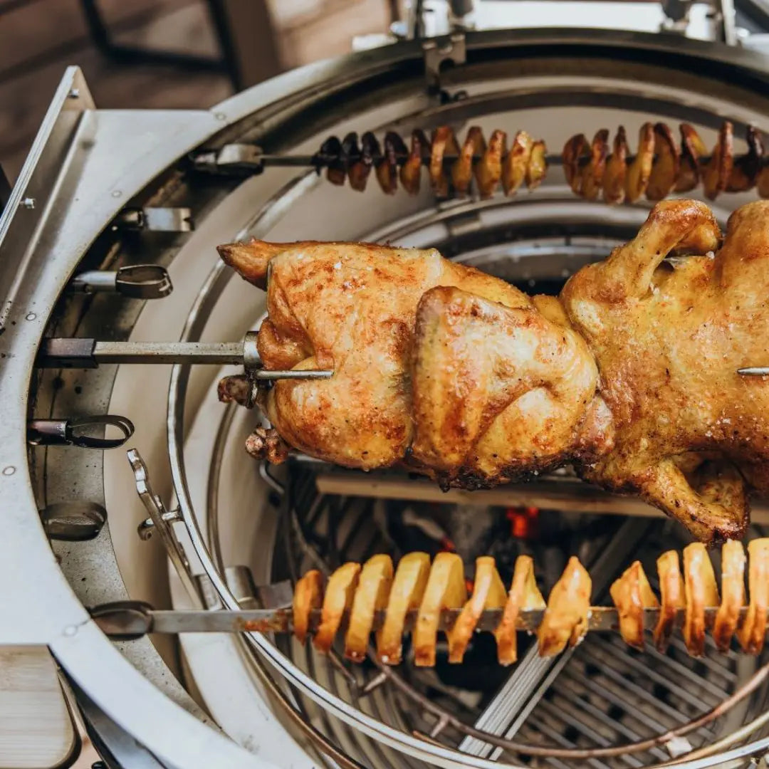 A close-up view of Monolith .55 rotisserie grill cooking a whole chicken on a metal spit, with sliced potatoes on another spit inside the grill.