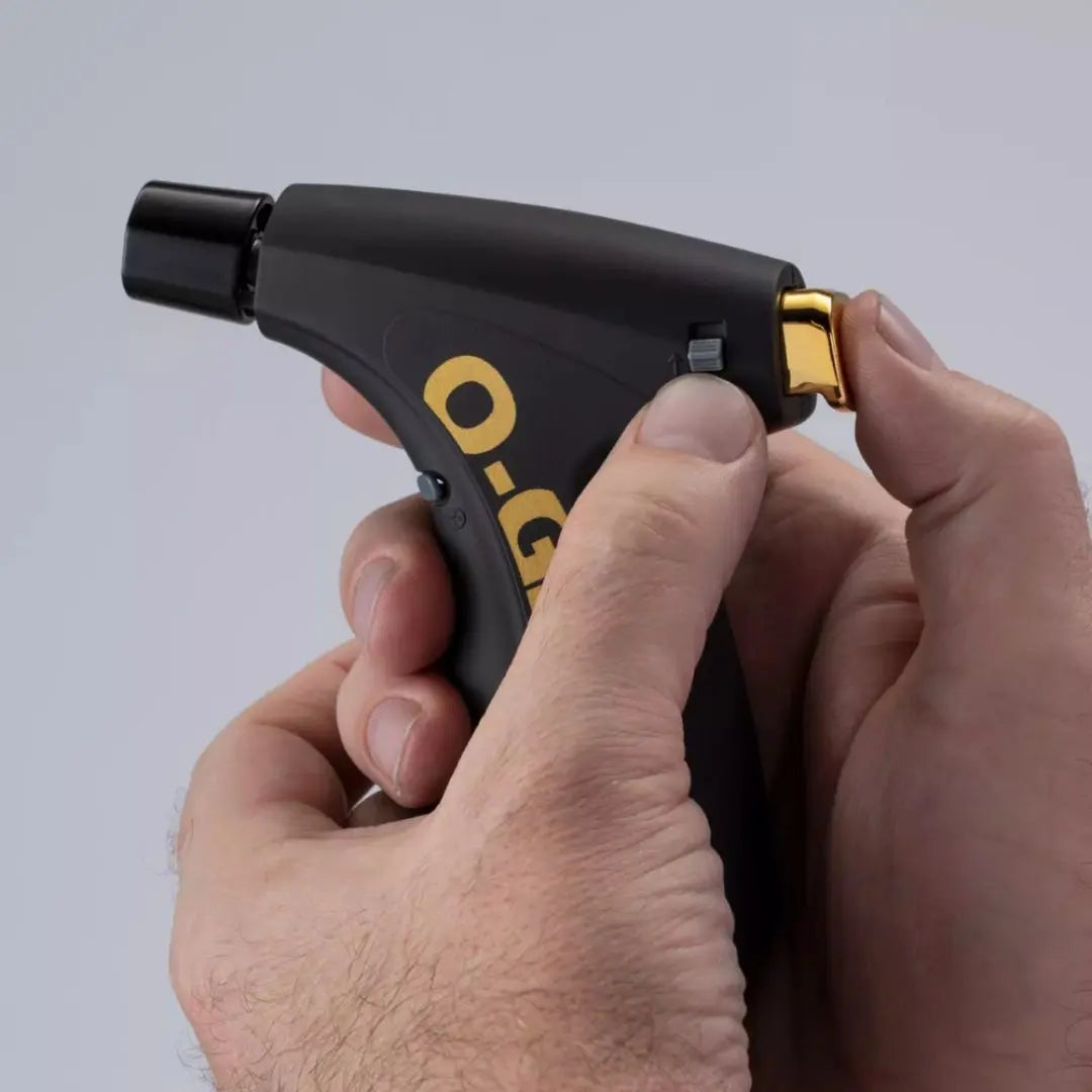 Hand holding a black and gold O-Grill torch lighter against a grey background