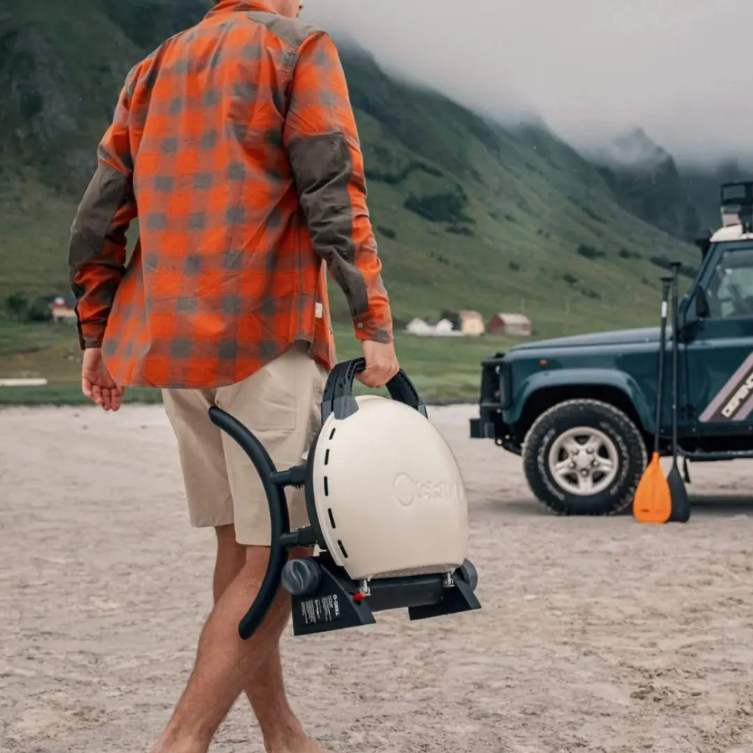 Person in orange plaid shirt carrying O-Grill portable gas grill bbq with Land Rover in a mountainous area.