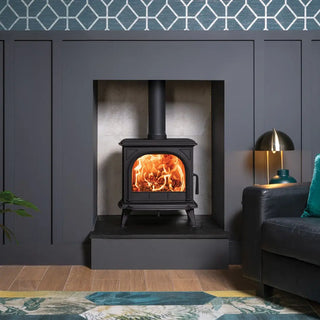 Stovax Huntingdon 30 Wood stove with clear doorin a room with a grey wall, sofa, and decorative elements.