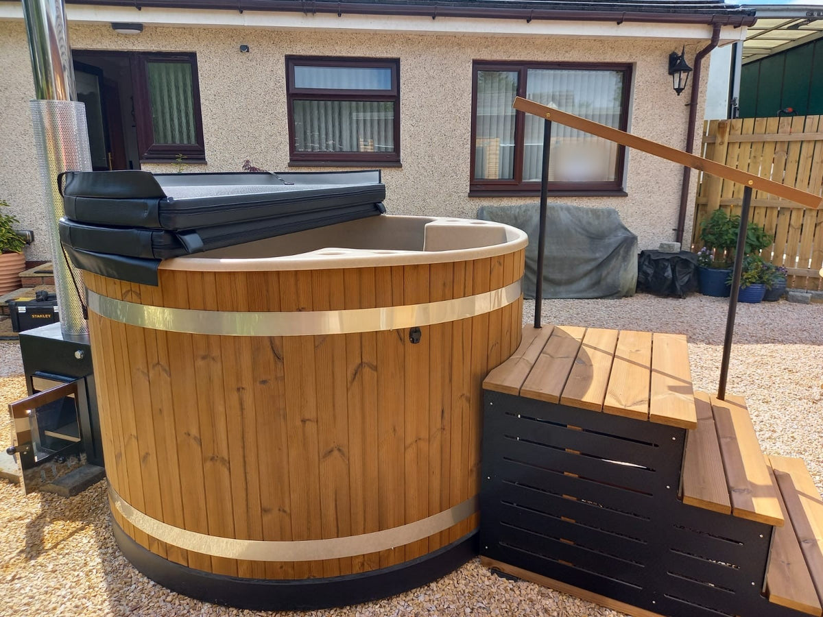 A round wooden hot tub with a wood-fired chimney, wooden steps and its lid folded back, in a gravel back garden