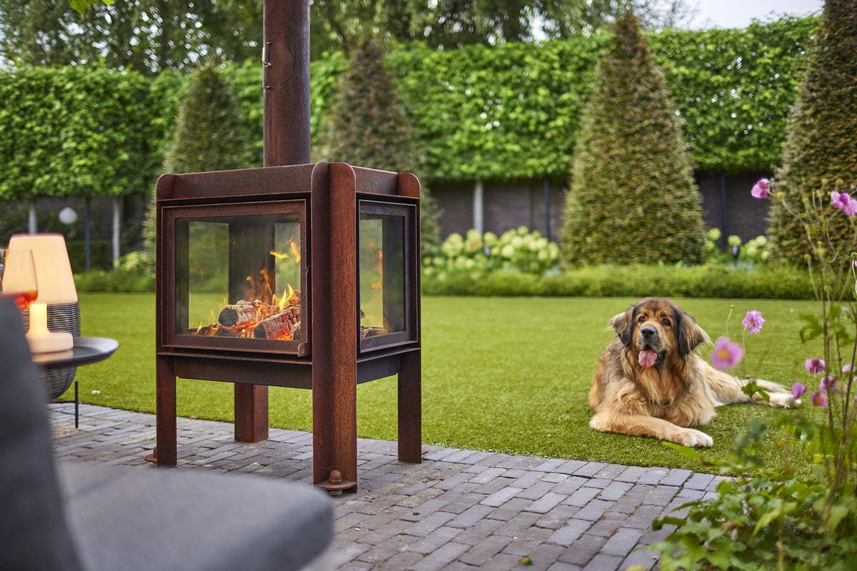 A lit RB73 outdoor fireplace in rust-coloured CorTen steel on a garden patio, with a dog lying on the lawn behind