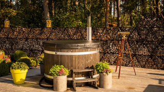 Wooden hot tub on a wooden deck with potted plants and stacked firewood in the background.